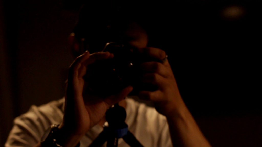 A darkly lit figure is taking aim towards you with their camera, ready to snap a photo.